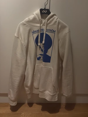 Vit Håkan Hellström hoodie L - Vit hoodie med blått tryck av Håkan Hellström på bröstet. Klassisk känguruficka framtill och justerbar huva med snören. Tillverkad i mjukt bomullsmaterial som är skönt att ha på sig. Perfekt för dig som gillar svensk musik och vill ha en unik hoodie. Köpt på Ullevi 13 juni 2025. Aldrig använd. Kan sänka pris vid snabb affär