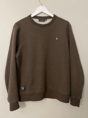 Brun sweatshirt från Morris - Säljer en brun sweatshirt från Morris med rund hals och ribbade muddar. Tröjan har en liten broderad logotyp på bröstet och en patch nertill. Perfekt för en avslappnad och stilren look.