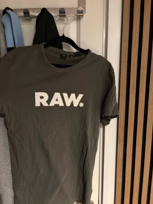 G-Star RAW grå t-shirt med tryck - Snygg grå t-shirt från G-Star RAW med stort vitt RAW-tryck på bröstet. Klassisk rund halsringning och korta ärmar. Perfekt för en avslappnad och trendig look. Tillverkad i mjuk bomull för skön komfort.