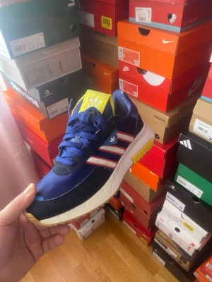 Adidas Retropy F2 blå sneakers med röda ränder - Snygga sneakers från Adidas med blå ovandel, vita och röda klassiska stripes på sidorna samt gula detaljer på plösen och sulan. Skon har en sportig siluett med vit mellansula och gummiyttersula. Perfekt för dig som gillar streetstyle och vill sticka ut.