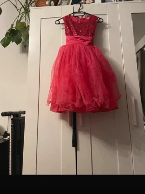 Röd glittrig prinsessklänning - Snygg röd prinsessklänning med glittrigt liv och fluffig tyllkjol. Klänningen har ärmlös design, markerad midja med satinband och stor rosett bak. Perfekt för speciella tillfällen när du vill sticka ut. Det är till barn runt 5 år