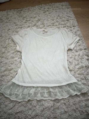 Vit topp med spetskant från H&M - Söt vit topp från H&M med korta ärmar och rund halsringning. Nederdelen har en bred, transparent spetskant med broderade detaljer som ger en romantisk touch. Perfekt att styla med jeans eller kjol.