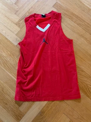 Röd basketlinne från Jordan - Snyggt rött basketlinne från Jordan med svart Jumpman-logga på bröstet och vit detalj vid halsen. Ärmlös modell med sportig passform, perfekt för träning eller match. Tillverkad i lätt och ventilerande material.
