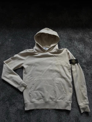 Beige hoodie från Stone Island - Snygg beige hoodie från Stone Island med klassisk patch på vänster ärm. Hoodien har känguruficka framtill och ribbade muddar vid ärmslut och nederkant. Perfekt för en avslappnad och stilren look.