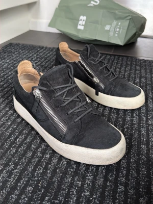 Giuseppe Zanotti skor - Svarta sneakers i mocka från Giuseppe Zanotti med silverfärgade dragkedjor på sidorna och vita, tjocka sulor. Skorna har svarta skosnören och en låg profil. Insidan är ljusbeige och materialet känns lyxigt och trendigt. Storlek 42
