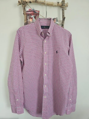 Rutig skjorta från Ralph Lauren - Säljer en klassisk långärmad skjorta från Ralph Lauren med rosa och vita rutor. Skjortan har knappar framtill, button-down krage och det ikoniska broderade logotypen på bröstet. Perfekt för dig som gillar stilrena och tidlösa plagg.