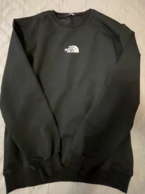 Svart sweatshirt från The North Face - En stilren svart sweatshirt från The North Face med klassisk rund halsringning och vit broderad logga på bröstet. Tröjan har ribbade muddar vid ärmslut och nederkant. Perfekt för en avslappnad och trendig look.