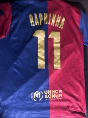 Barça matchtröja Raphinha #11 - Säljer en officiell FC Barcelona matchtröja med Raphinha och nummer 11 på ryggen. Tröjan är blå och röd med gul text, Spotify-logga på framsidan och UNHCR ACNUR på baksidan. Materialet är lätt och andas, perfekt för fotboll eller träning.