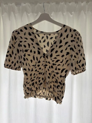 Beige blus med svart mönster - Trendig beige blus med svart abstrakt mönster och v-ringning. Blusen har korta puffärmar och rynkdetalj framtill som ger en snygg siluett. Tillverkad i ett lätt och luftigt material, perfekt för varma dagar.