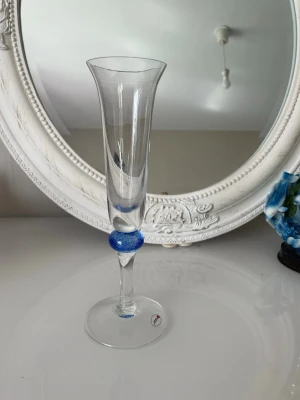 Elegant blå detalj glas  - Säljer en smal och hög glas med en blå dekorativ detalj på foten. Vasen är transparent med en blå ton i botten och har en böjd form upptill. Perfekt som dekoration eller för enstaka blommor.