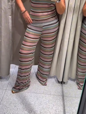Randiga vida leggings i pastell - Säljer ett par vida leggings med horisontella ränder i pastellfärger som rosa, grön, blå, beige och svart. De har en bred resår i midjan och är tillverkade i ett tunt, stretchigt material som ger en cool och avslappnad look. Några små trådar som sticker ut