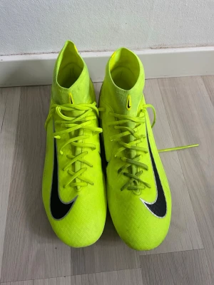 Nike Mercurial gula fotbollsskor - Säljer ett par Nike Mercurial fotbollsskor i neon-gult med svarta detaljer och högt skaft. Skorna har snörning och slitstarka dobbar för bra grepp på planen. Tillverkade i syntetmaterial för lätt känsla och snabbhet. Perfekta för dig som vill sticka ut på fotbollsplanen.