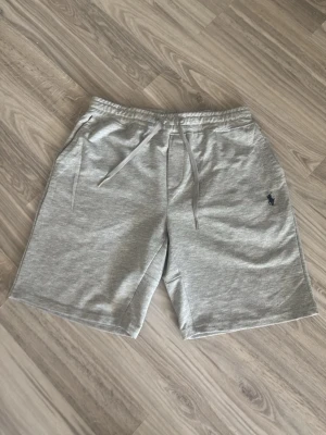 Grå shorts från Polo Ralph Lauren - Snygga grå shorts från Polo Ralph Lauren med klassisk logga broderad på vänster ben. Modellen har dragsko i midjan och två sidofickor. Perfekta för en avslappnad stil och tillverkade i mjuk bomull för extra komfort.