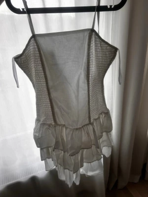 Vit kort klänning med volanger perfekt till studenten! - Söt vit kort klänning med smala axelband och smockad överdel. Klänningen har volangdetaljer nedtill och är tillverkad i ett lätt, luftigt tyg som passar perfekt för varma dagar. Enkel och stilren design med romantisk känsla. Väldigt fin klänning till studenten! 