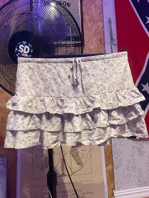 Blommig volangkjol med snörning - Supersöt kort kjol med blommigt mönster i ljusa toner. Kjolen har flera volanger och en snörning i midjan för extra detalj. Perfekt för dig som gillar romantisk och feminin stil.