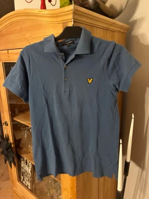 Blå pikétröja från Lyle & Scott - Klassisk blå pikétröja från Lyle & Scott med korta ärmar och gul logga på bröstet. Tröjan har krage och knappar framtill. Tillverkad i mjuk bomull som känns skön mot huden. Perfekt för en avslappnad och stilren look. Använd ett fåtal gånger och har storleken M
