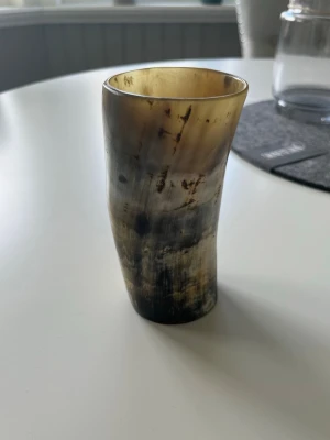 Dryckeshorn i polerat horn - Unikt dryckeshorn från Nordlys, tillverkat i polerat horn med naturliga färgskiftningar i brunt, svart och beige. Formen är avsmalnande och botten är försedd med en träplatta med inbränt märke. Den kan va till för att dricka ur den men även ha som prydnad. Den är helt ny och har aldrig använts, kvitto finns med på den. Hör av er om ni har frågor!
