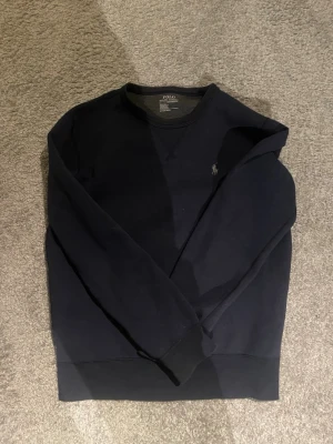 Mörkblå sweatshirt från Polo Ralph Lauren - Klassisk mörkblå sweatshirt från Polo Ralph Lauren med rund halsringning och diskret broderad logga på bröstet. Tillverkad i mjukt bomullsmaterial och har ribbade muddar vid ärmslut och nederkant. Perfekt för en avslappnad och stilren look.