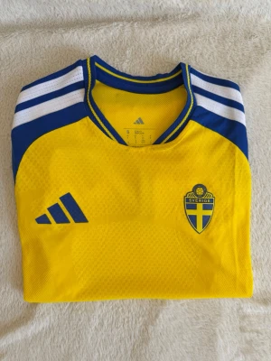 Sverige fotbollströja VM2026 - Gul och blå fotbollströja från Adidas med Sveriges landslagsemblem på bröstet. Tröjan har vita och blå ränder på axlarna, rund hals och är tillverkad i lätt och ventilerande material med ClimaCool-teknologi. Perfekt för träning eller match. Sitter mer som en XS på killar men som S på tjejer! (Killmodell)