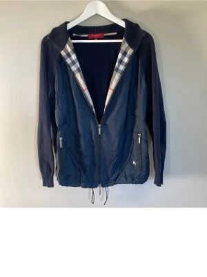 Burberry cardigan  - Snygg marinblå cardigan från Burberry med klassiskt rutigt foder i huvan. Jackan har dragkedja framtill, två fickor med dragkedja och ribbade muddar på ärmarna. Perfekt för dig som vill ha en stilren och tidlös look.