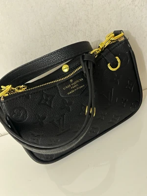 Svart Louis Vuitton axelväska i skinn - Säljer en svart axelväska från Louis Vuitton med klassiskt monogrammönster i präglat skinn. Väskan har guldfärgade metalldetaljer, dragkedja och både axelrem i skinn och kedja. Perfekt storlek för det viktigaste och riktigt snygg design.