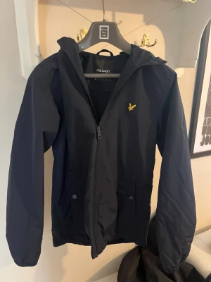 Marinblå vindjacka från Lyle & Scott - Stilren marinblå vindjacka från Lyle & Scott med dragkedja framtill, huva och två framfickor med tryckknappar. Är tillverkad i ett lätt, vindtätt material. Perfekt för blåsiga dagar. 
