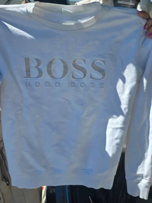 Vit sweatshirt från Hugo Boss - Säljer en stilren vit sweatshirt från Hugo Boss med stor BOSS-logga broderad framtill. Tröjan har rund halsringning och långa ärmar. Perfekt för dig som gillar klassisk och enkel design med premiumkänsla.