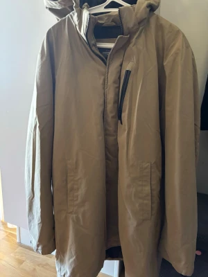 Beige dunjacka från dressman  - Snygg beige dunjacka från dressman med huva och dragkedja. Jackan har en stilren design med två sidofickor och en bröstficka med svart dragkedja. Perfekt för kalla vinterdagar och enkel att matcha med olika outfits.