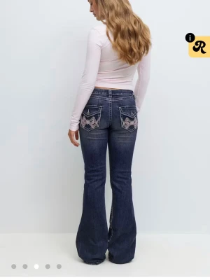 iconic jeans  - snygga mörkblå bootcut jeans med slitningar och broderade bakfickor i rosa och vitt. Jeansen har dekorativa sömmar och ficklock med knapp. köpa på Gina Young