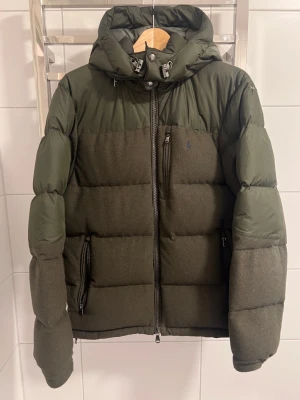 Ralph lauren jacka - Mörkgrön pufferjacka från Polo Ralph Lauren med huva och dragkedja framtill. Jackan har quiltad design, två sidofickor med dragkedja och en bröstficka. Materialmix med glansiga och matta partier samt diskret logga på bröstet.