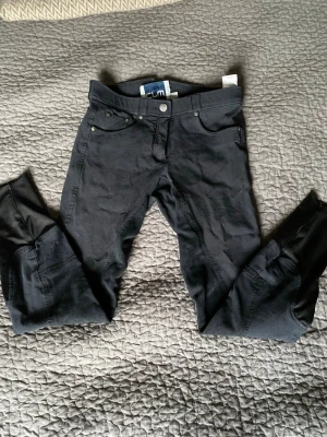 Svarta ridbyxor från Crw - Snygga svarta ridbyxor från Crw med klassisk femficksdesign och förstärkta partier på insidan av benen. Byxorna har normal midja och bälteshällor, samt knäppning med knapp och dragkedja. Perfekta för dig som gillar ridning och vill ha en stilren look. En liten söm har släppt vid knäppningen, men som inte är så synlig.