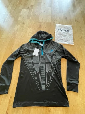 Svart hoodie från Gymshark med mönster - Svart hoodie från Gymshark med grått geometriskt mönster och turkosa detaljer vid huvan. Långärmad modell i ett stretchigt, sportigt material som passar perfekt till träning. Gymshark-logga i turkos på bröstet och dragkedja vid halsen.