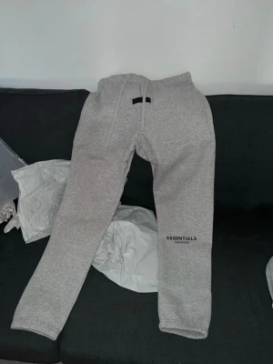 Grå Essentials Fear of God mjukisbyxor - Snygga grå mjukisbyxor från Essentials Fear of God med logga tryckt på benet. Byxorna har resår i midjan och benslut samt dragsko för bästa passform. Perfekta för en avslappnad och trendig look.