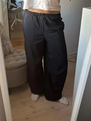 Svarta baggy cargobyxor  - Säljer ett par svarta cargobyxor med bred passform och elastisk midja. Byxorna har stora fickor på sidorna och är tillverkade i ett lätt, glansigt material som ger en cool streetkänsla. Perfekta för en avslappnad och trendig look.