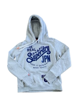 Grå hoodie från Superdry med tryck - Snygg ljusgrå hoodie från Superdry med stort blått tryck på bröstet och japanska tecken. Klassisk känguruficka framtill och justerbar huva med dragsko. Perfekt för en avslappnad streetstyle-look. Är storlek M men sitter som S
