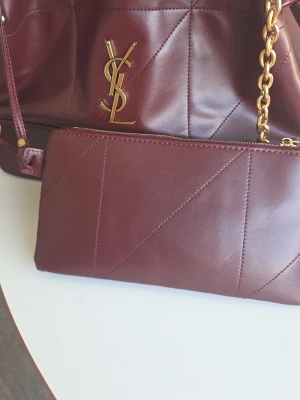 Vinröd YSL handväska i läder - Snygg vinröd handväska från YSL i mjukt läder med guldfärgade kedjedetaljer och ikoniska YSL-loggan framtill. Väskan har quiltade sömmar och en matchande clutch med dragkedja. Perfekt accessoar för dig som vill sticka ut med stil.