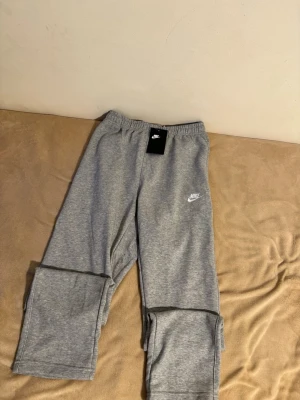 Grå Nike mjukisbyxor med logga - Snygga grå mjukisbyxor från Nike med broderad vit logga på benet. Byxorna har elastisk midja och är tillverkade i mjukt och bekvämt material som passar perfekt för chill eller träning. Klassisk rak passform och stilren design.