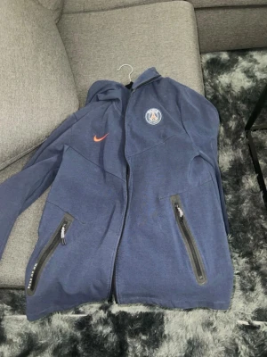 Blå PSG hoodie från Nike - Säljer en blå hoodie från Nike med PSG-logga på bröstet och orange Nike-swoosh. Hoodien har dragkedja framtill, två fickor med dragkedja och huva. Perfekt för dig som gillar sport och streetwear. Materialet är mjukt och bekvämt.