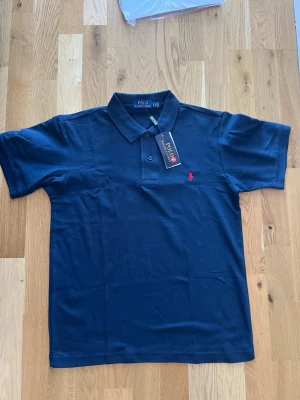 Marinblå pikétröja från Polo Ralph Lauren - Klassisk marinblå pikétröja från Polo Ralph Lauren med korta ärmar och krage. Tröjan har knappar framtill och den ikoniska röda logotypen broderad på bröstet. Perfekt till jeans eller shorts för en clean look.