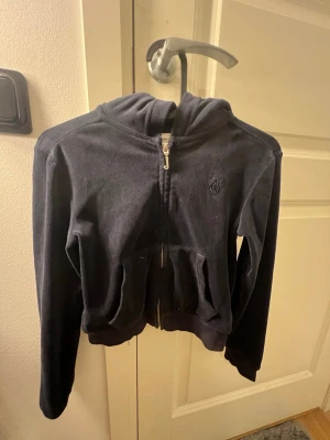Mörkblå velour hoodie från Juicy Couture - Säljer en mörkblå hoodie i mjuk velour från Juicy Couture. Tröjan har dragkedja framtill, två fickor och en broderad logga på bröstet. Klassisk huva och ribbade muddar vid ärmslut och nederkant. Perfekt för en chill och trendig look.