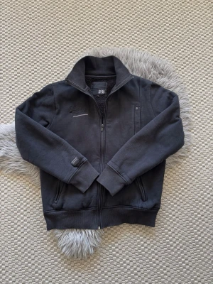 Svart fleecejacka från Jack & Jones - Svart fleecejacka från Jack & Jones med dragkedja framtill, hög krage och ribbade muddar. Jackan har två sidofickor med dragkedja och en liten bröstficka. Perfekt för kyliga dagar och enkel att matcha med olika outfits.