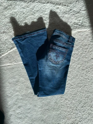 Blå bootcut jeans med broderad ficka - Snygga blå bootcut jeans med broderad bakficka och röda sömmar. Jeansen har klassisk femficksdesign. Har små kvarg jeans detaljer typ. Perfekta för dig som gillar en retroinspirerad look och vill sticka ut med coola detaljer.