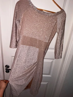 Glittrig beige långklänning - Säljer en glittrig beige långklänning med trekvartsärm och slits framtill. Klänningen har en snygg omlottkänsla och är perfekt för dig som vill sticka ut. Materialet är mjukt och stretchigt, vilket gör den bekväm att bära hela kvällen.
