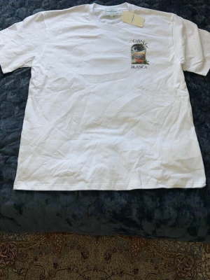 Vit Casa Blanca t-shirt med tryck - Vit t-shirt från Casablanca med tryck av ett fönster med palmer och stadsmotiv på ryggen samt mindre tryck på bröstet. Klassisk passform och rund halsringning. Perfekt för en chill och stilren look. Helt stiligt om du vill sticka ut under sommaren