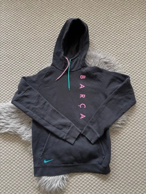 Svart Barça hoodie från Nike - Svart hoodie från Nike med vertikal rosa 'BARÇA'-text på bröstet och färgglada dragsnören i rosa och turkos. Klassisk känguruficka och Nike-logga i turkos vid midjan. Perfekt för dig som gillar sportig och stilren streetwear.