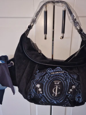 Svart axelväska från Juicy Couture - Cool svart axelväska från Juicy Couture med broderad logga och texten Los Angeles framtill. Väskan har blått och vitt mönster, samt en blå och mörkblå scarf knuten i sidan. Mjuk mockaliknande yta och metalliska detaljer på handtaget.