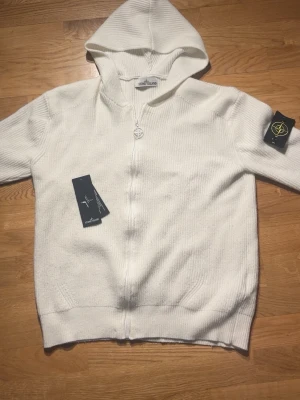 Stone island knitted zip up hoodie - Vit stickad hoodie från Stone Island med dragkedja framtill och klassisk logotyp-patch på ärmen. Tröjan har ribbade muddar och är tillverkad i ett mjukt, strukturerat material. Perfekt för en clean och stilren look.