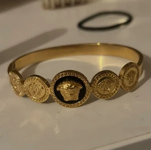  Versace armband - Snyggt armband i guldfärg från Versace med flera runda plattor och ikoniska Medusa-huvuden. Armbandet har en lyxig känsla med detaljer i svart och guld.