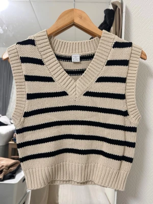 Randig stickad väst från Lindex - Beige och svart stickad väst från Lindex med bred v-ringning och ribbade kanter. Västen har ett klassiskt randigt mönster och är perfekt att styla över en skjorta eller t-shirt för en trendig look.