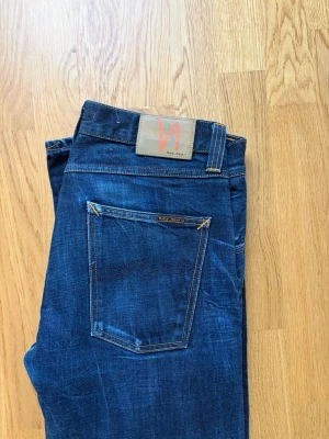 Mörkblå jeans från Nudie Jeans - Säljer ett par mörkblå jeans från Nudie Jeans med klassisk femficksmodell och raka ben. Jeansen har kontrastsömmar och en snygg läderpatch med logga bak i midjan. Perfekta för dig som gillar stilrena och tidlösa jeans i slitstarkt denimtyg.